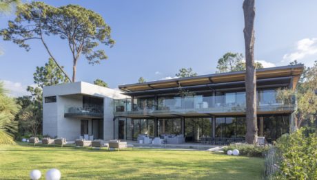 Modern two-story house with glass walls surrounded by trees.