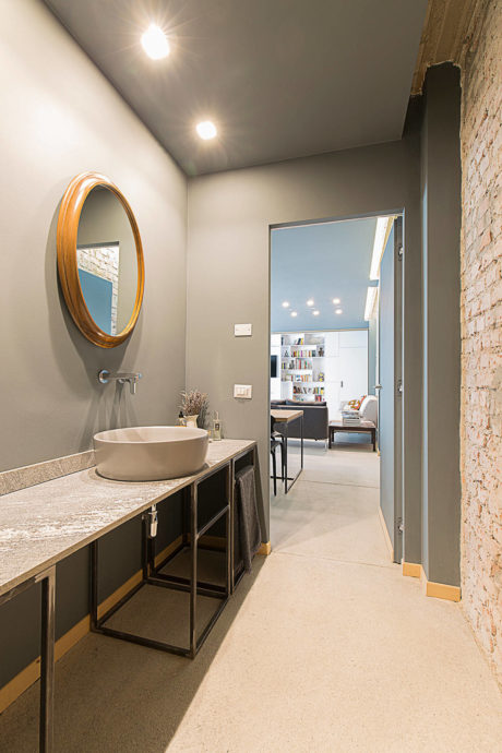 Modern bathroom with oval mirror, exposed brick, and view into living space.