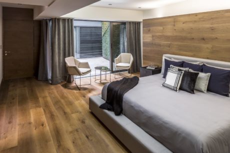 Modern bedroom with wooden flooring, large bed, and seating area leading to a patio