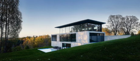 Modern glass-walled house with flat roof on a grassy slope at dusk.