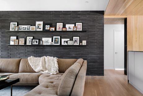 Modern living room with black brick wall and photo frames above a tan couch.
