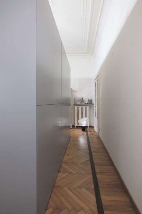 Narrow hallway with herringbone flooring and white walls leading to a bright room