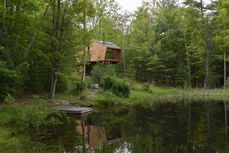 Treehouse with large windows overlooking a tranquil pond in a wooded area.