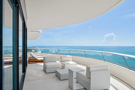 Modern oceanfront balcony with white furniture and glass railing.