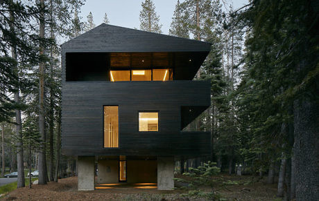 Angular black-clad house with lit windows amidst a forest.