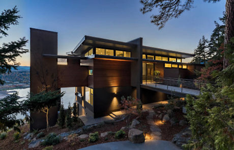 Modern hillside house with large windows at dusk overlooking a body of water.