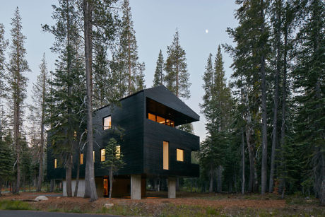 Modern black cabin in a forest with lights on at dusk.