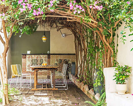 Lush greenery frames a cozy outdoor nook with a rustic table and chairs, patterned tiles.