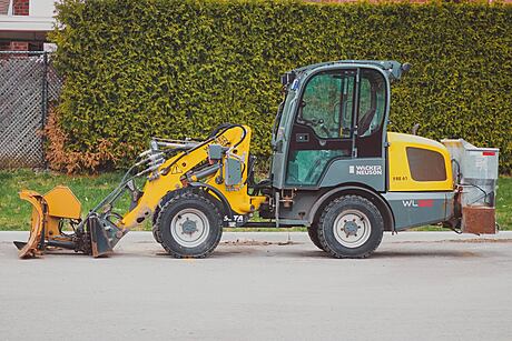 A yellow construction vehicle with a forklift attachment is parked in front of a brick building with lush greenery.