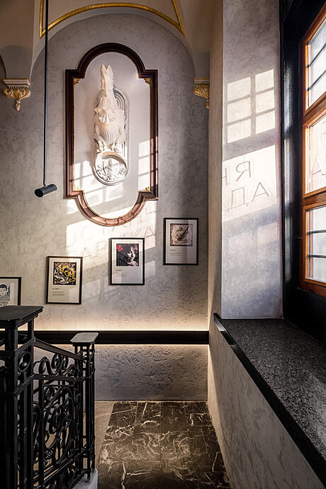 A grand, ornate archway frames a sculpted statue against a marble backdrop, with decorative lighting and framed artwork.