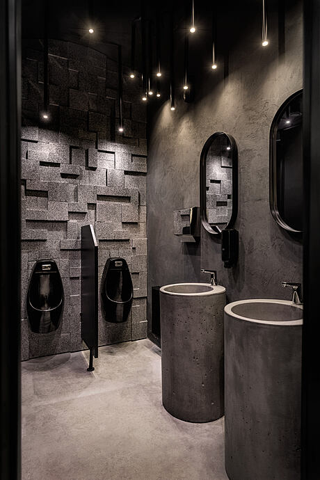 Dark, modern bathroom with concrete sinks, tiled walls, and hanging light fixtures.