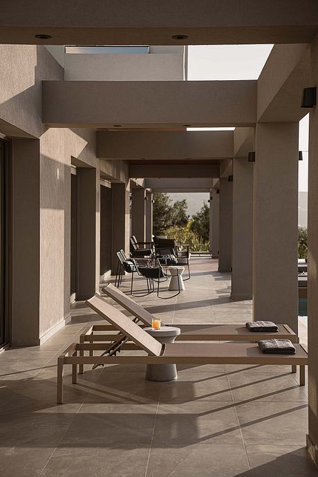 A minimalist outdoor patio with chic lounge chairs, a low table, and a tiled floor leading to a covered walkway.