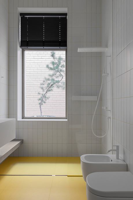 Minimalist bathroom with sleek white tiles, yellow floor, and a window offering a glimpse of nature.
