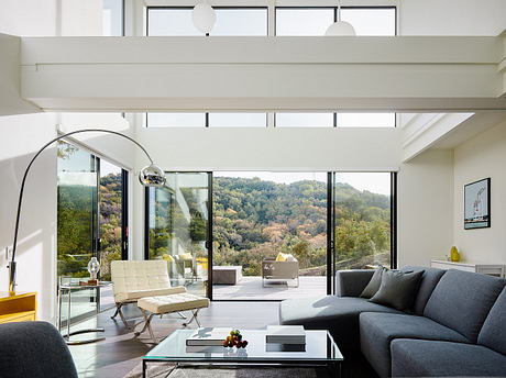 A modern, minimalist living room with large windows overlooking a lush, forested landscape.