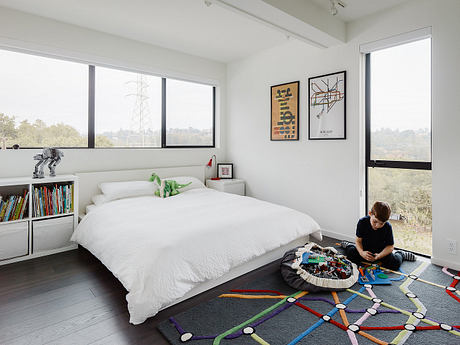 A modern bedroom with large windows, bookshelves, and a colorful patterned rug.