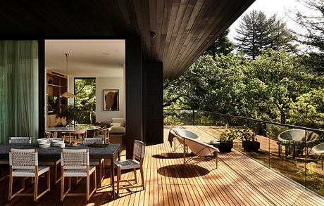 Spacious indoor-outdoor living area with wooden deck, glass railing, and lush greenery.
