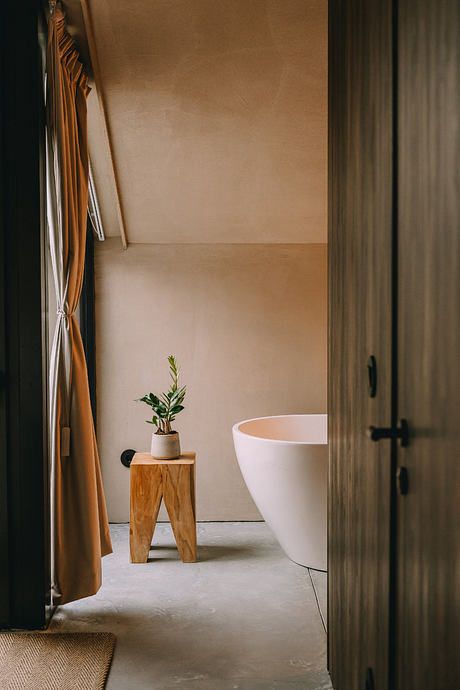 A cozy bathroom with a modern bathtub, wooden stool, and a potted plant, set in a warm, minimalist space.