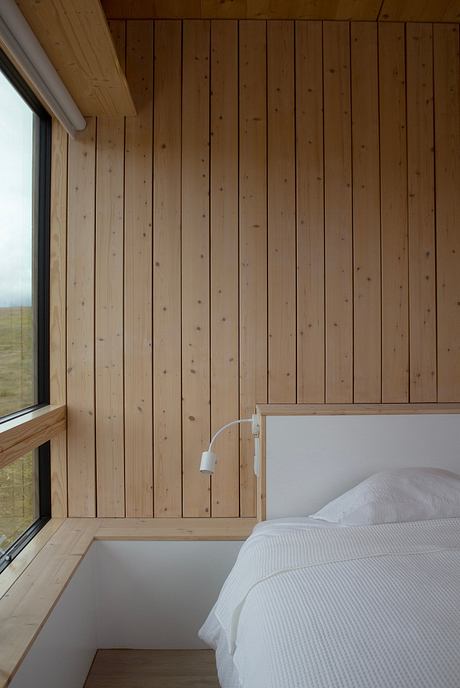 A cozy bedroom with wooden paneled walls, a large window, and a minimalist bed frame.