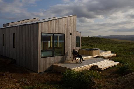 A modern wooden cabin nestled in a lush green landscape, with a deck and hot tub.