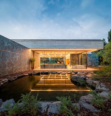 A modern, glass-walled structure with a stone wall and a pond reflecting its warm, inviting interior.