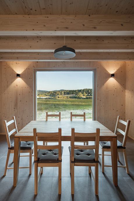 Warm wooden interior with modern dining table, chairs, and large window overlooking scenic landscape.