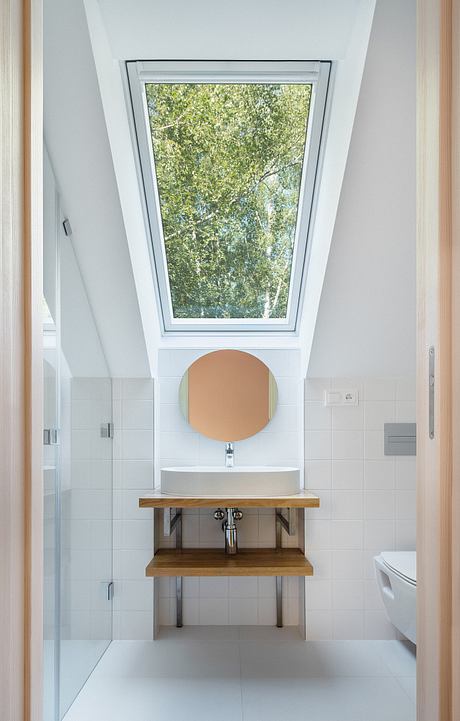 A modern bathroom with a circular mirror, wooden vanity, and large skylight window overlooking trees.