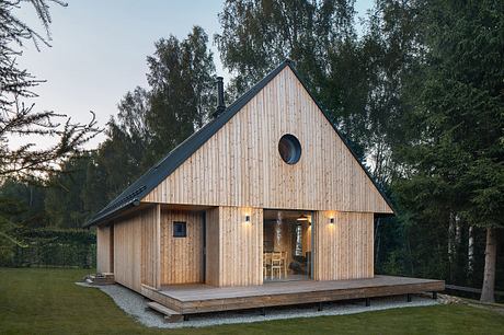 A rustic, wooden cabin nestled in a forest, featuring an asymmetric roof and a deck.