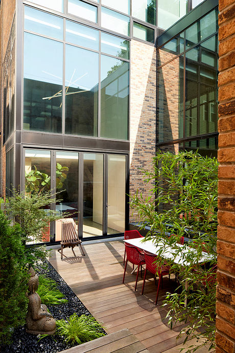 A modern glass-enclosed courtyard with a wooden deck, lush greenery, and red chairs.