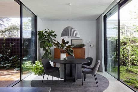 A modern, minimalist dining area with a circular wooden table, dark chairs, and lush plants.