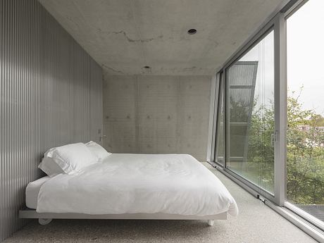 A minimalist concrete bedroom with floor-to-ceiling windows overlooking greenery.