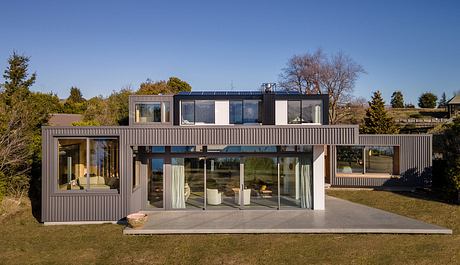 Modern, modular home with expansive windows, solar panels, and a concrete patio.