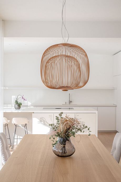 A modern, minimalist kitchen featuring a sculptural pendant light and a floral arrangement on a wooden table.