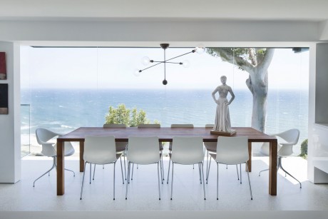 A modern, minimalist dining space with a large wooden table, white chairs, and a scenic ocean view.