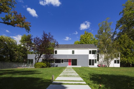 A modern, two-story white house with clean lines, a red front door, and surrounded by lush green trees.