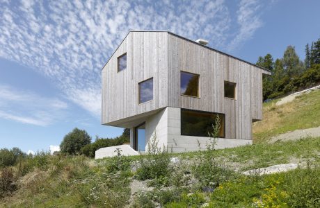 A modern, wooden house perched on a grassy hill, with large windows and a striking architectural design.