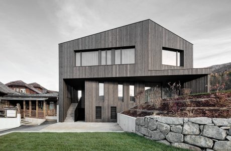 Modern, angular wooden structure with large windows, stone retaining wall, and grassy yard.