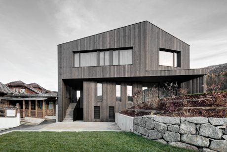 Modern, angular wooden structure with large windows, stone retaining wall, and grassy yard.