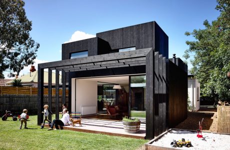 A modern, black-clad home with a large covered patio, wooden accents, and lush greenery.