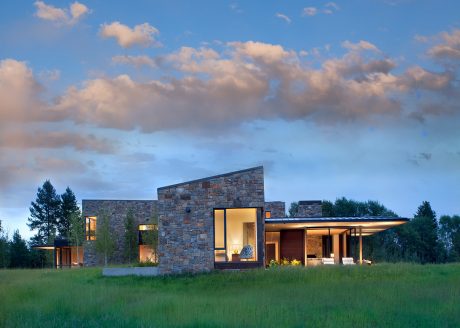 A modern, single-story home with stone exterior walls and large windows, set in a grassy field.