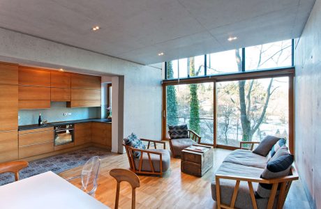 Modern, open-plan interior with concrete walls, wood cabinetry, and large windows overlooking trees.