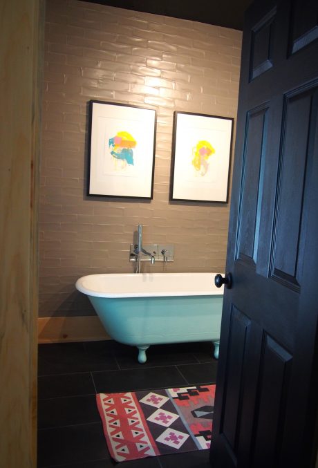 A cozy bathroom with a turquoise clawfoot tub, framed abstract art, and patterned tile floor.