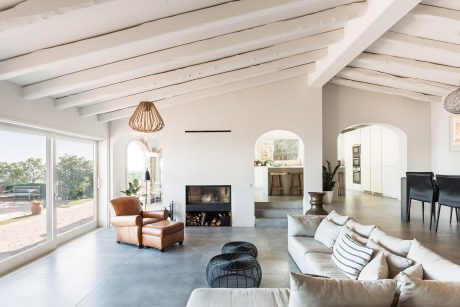 Modern, open-plan living room with exposed wooden beams, large windows, and a cozy fireplace.