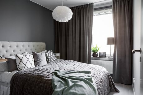 The image depicts a modern, cozy bedroom with a tufted gray headboard, plush bedding, and a stylish feather pendant light.