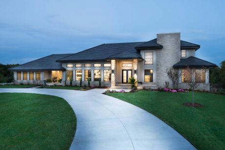 Large, modern multi-story home with stone exterior, slanted roof, and illuminated windows.