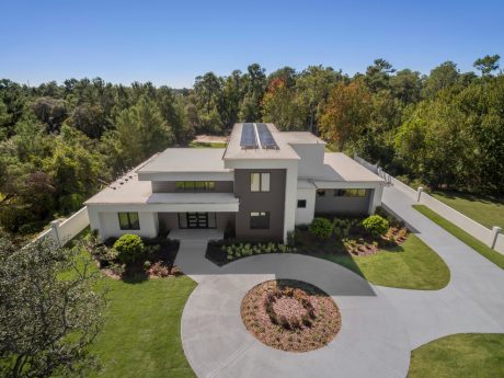 Modern single-story home with solar panels, white walls, and a circular driveway surrounded by lush greenery.