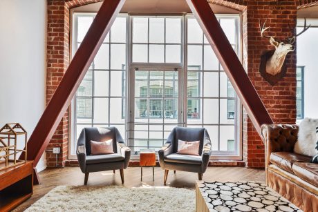 A loft-style room featuring brick walls, large windows, and wooden beams. Two armchairs sit on a plush rug, creating a cozy seating area.