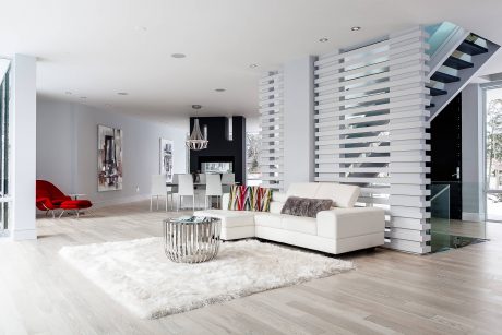 Modern, open-concept living room with sleek white furniture, glass staircase, and textured accents.