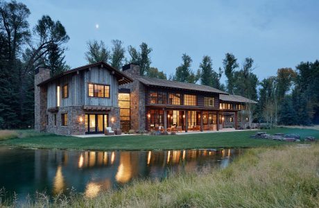 Rustic, multi-story wooden lodge with stone accents, surrounded by trees, reflecting on pond.