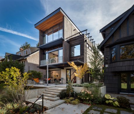 A modern two-story home with a mix of wood, glass, and stone elements, featuring a balcony and landscaped gardens.