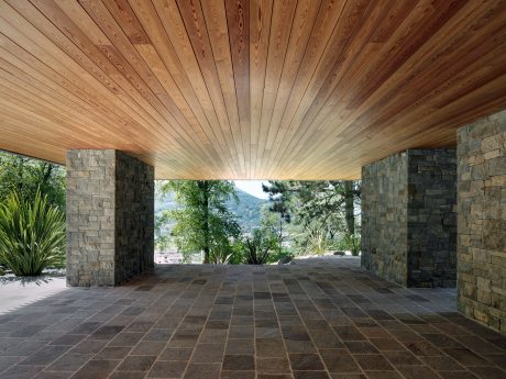 A covered porch with wood-paneled ceiling and stone walls, opening to a lush, forested landscape.